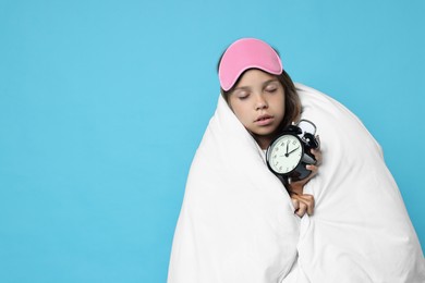 Overslept girl with sleep mask and alarm clock wrapped in blanket on light blue background, space for text