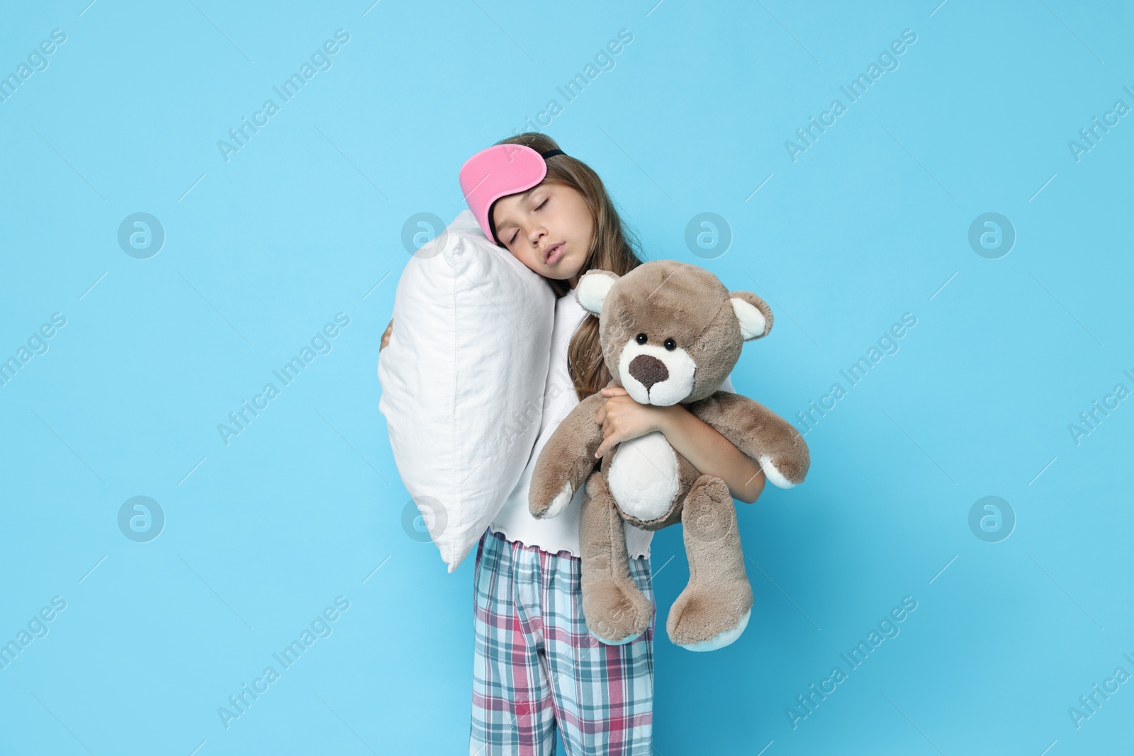 Photo of Overslept girl with sleep mask, pillow and teddy bear on light blue background