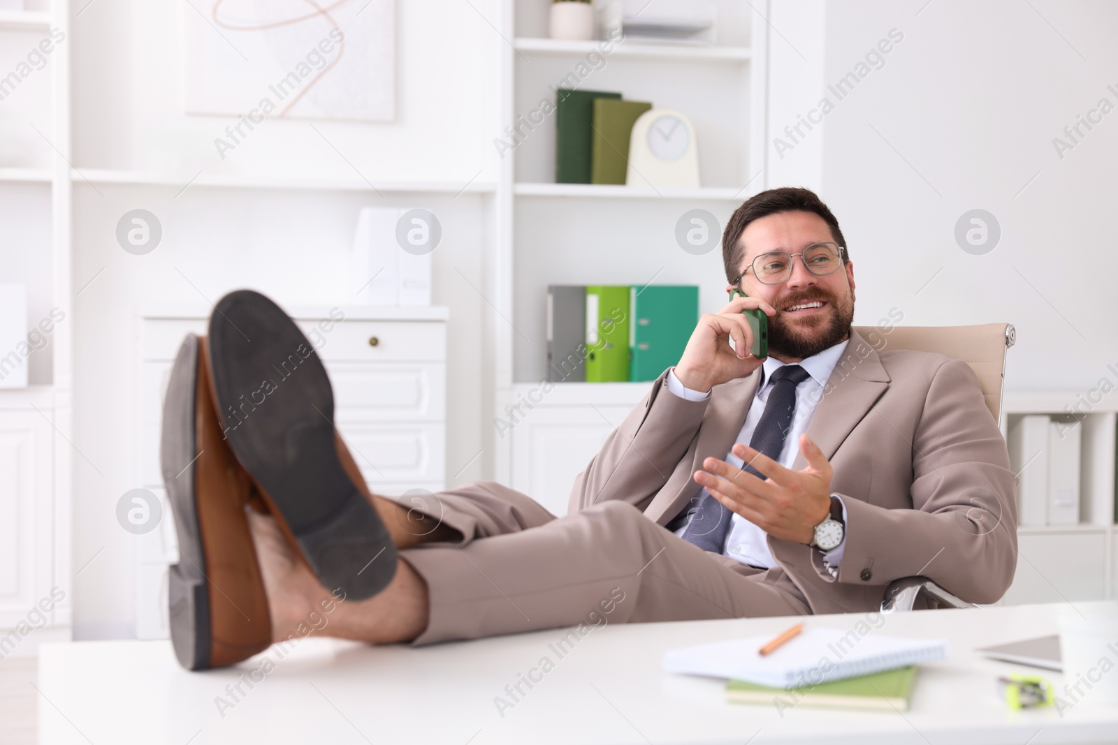 Photo of Smiling businessman talking by smartphone and holding legs on table in office