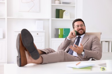 Smiling businessman talking by smartphone and holding legs on table in office