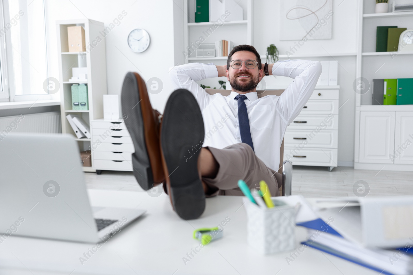 Photo of Smiling businessman with hands behind his head holding legs on table in office. Break time