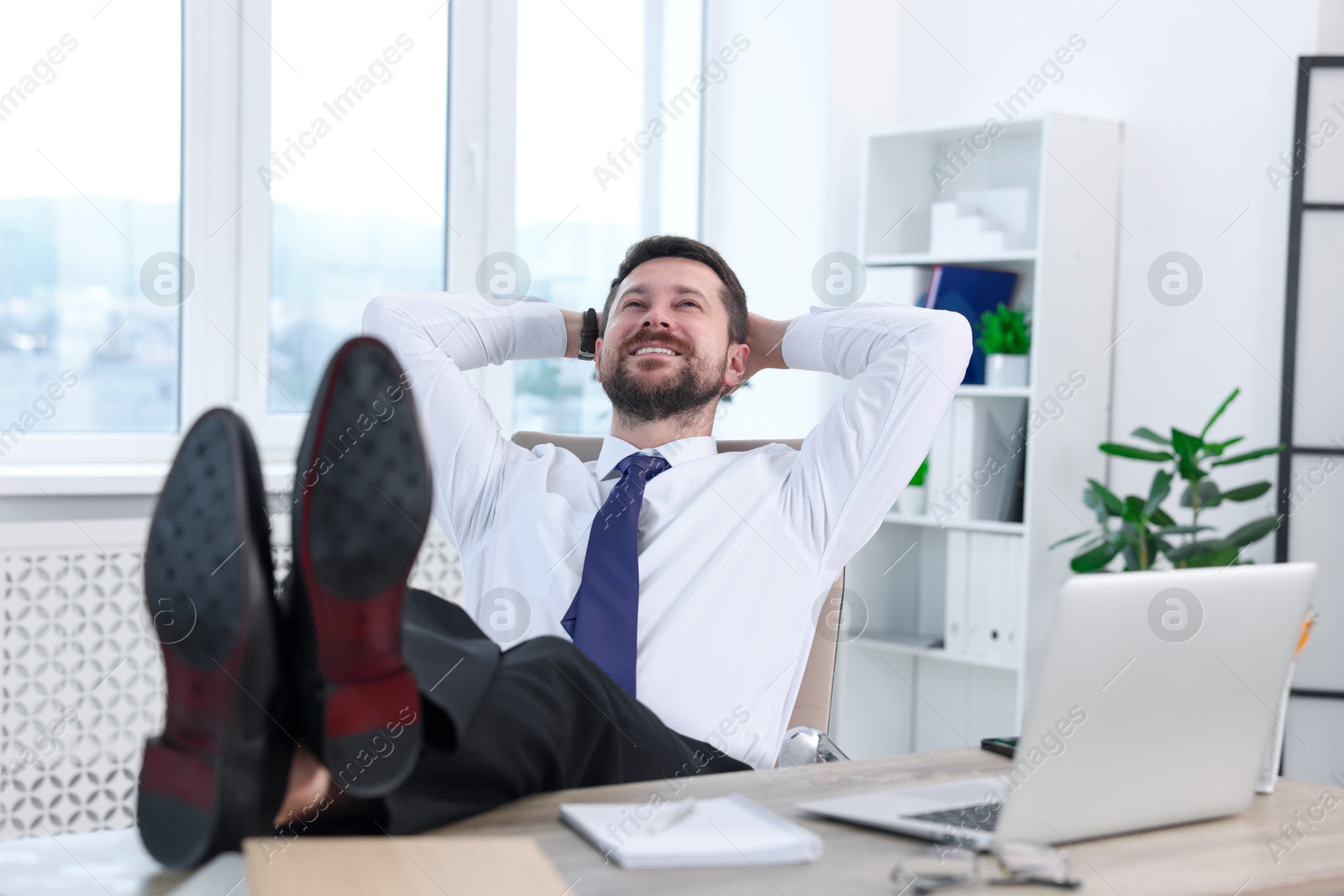 Photo of Smiling businessman with hands behind his head holding legs on table in office. Break time