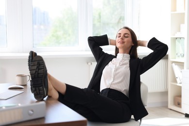 Photo of Smiling businesswoman with hands behind her head holding legs on table in office. Break time