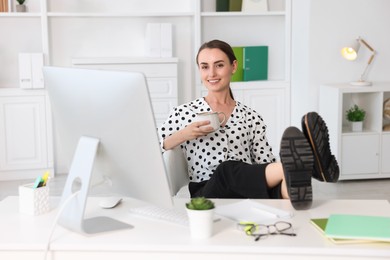 Photo of Smiling businesswoman drinking coffee holding legs on table in office. Break time