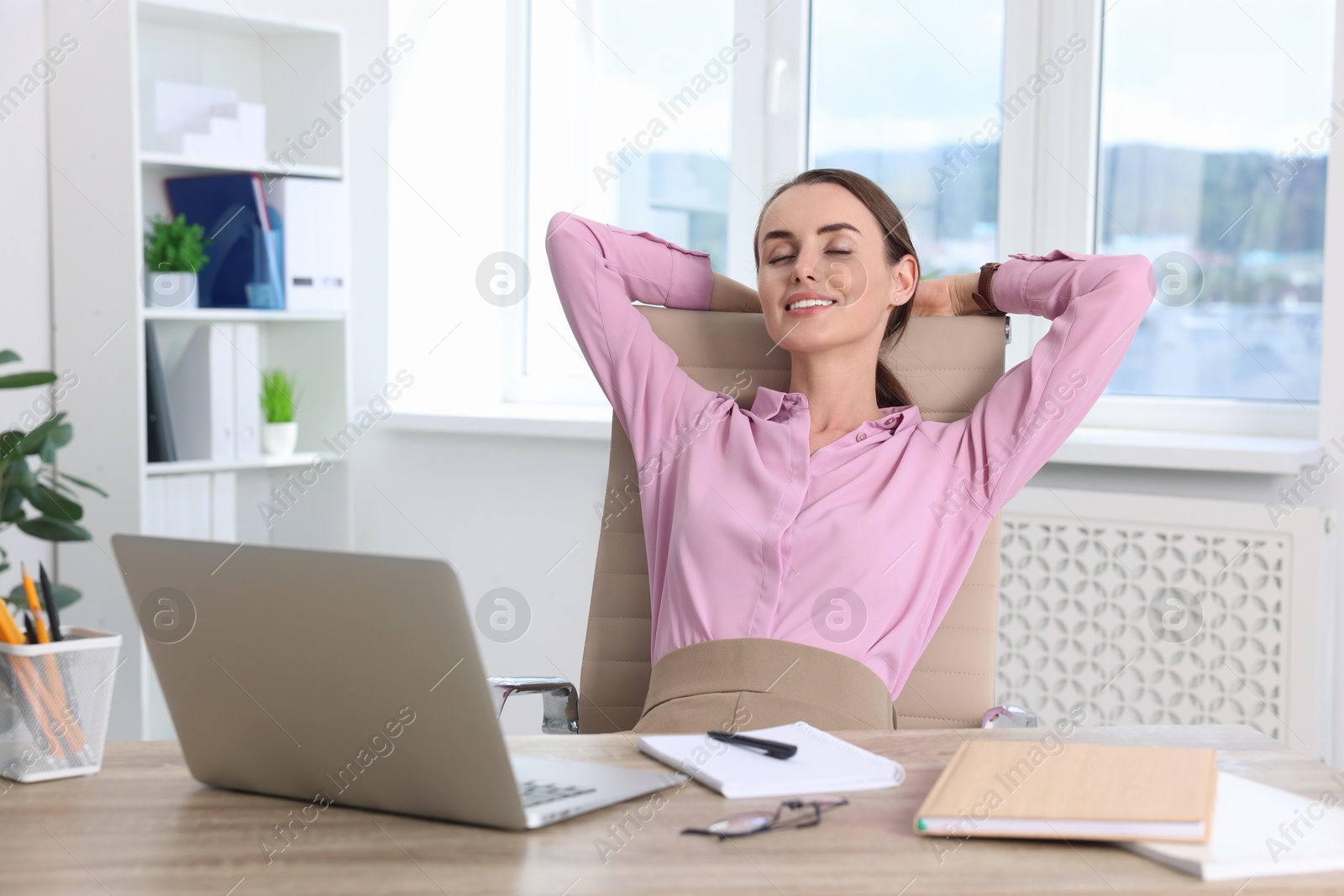 Photo of Smiling businesswoman with hands behind her head sitting at table in office. Break time