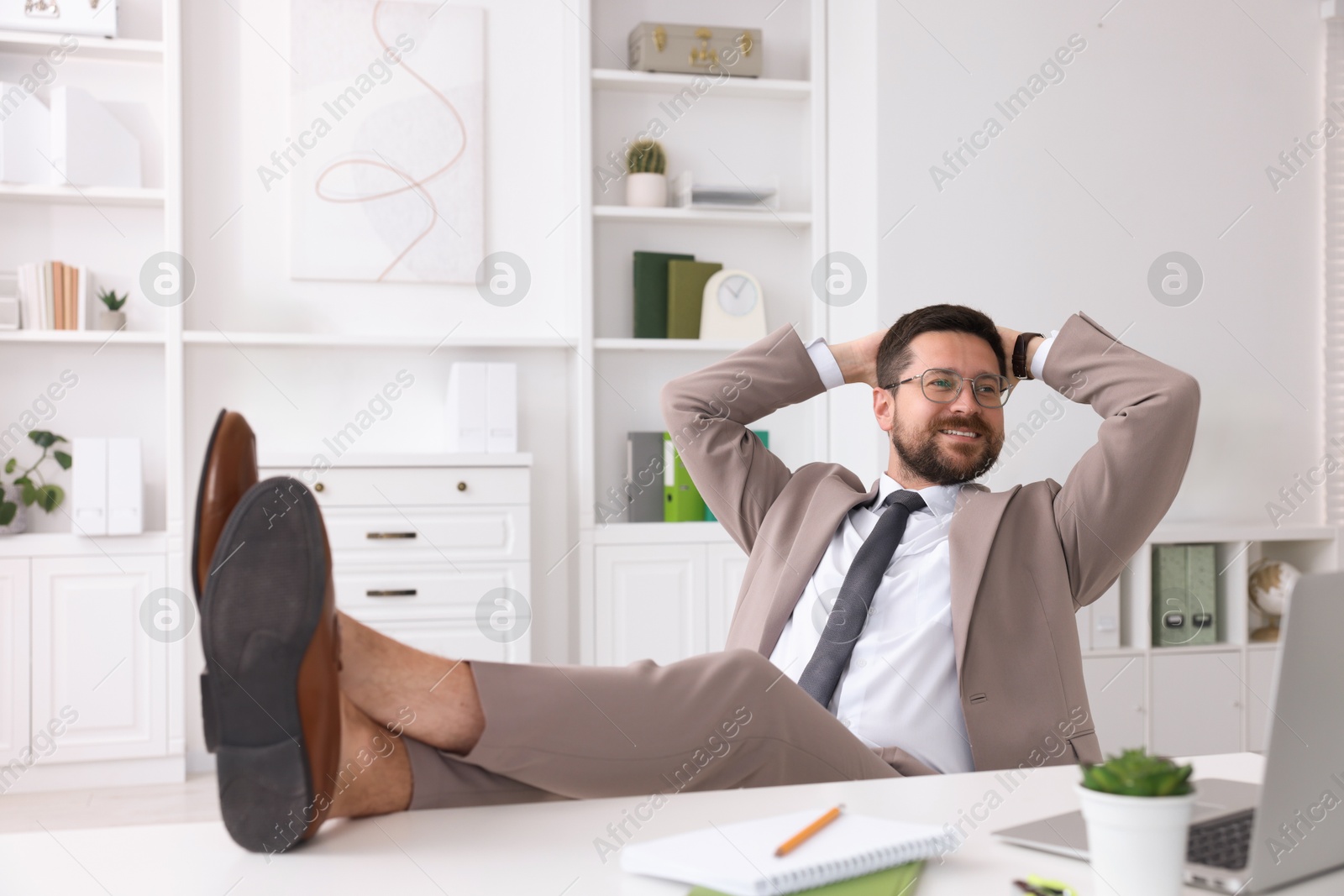 Photo of Smiling businessman with hands behind his head holding legs on table in office. Break time