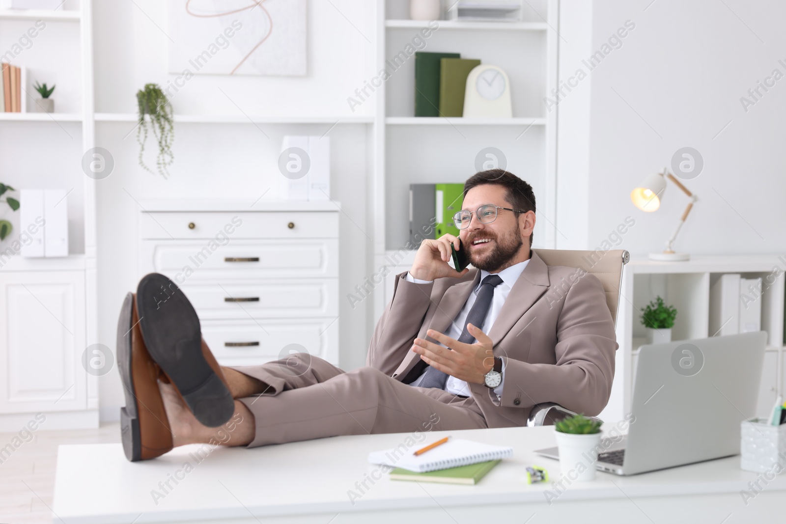 Photo of Smiling businessman talking by smartphone and holding legs on table in office