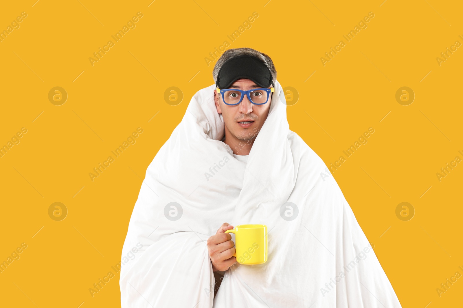 Photo of Overslept man with sleep mask, glasses and cup wrapped in blanket on orange background