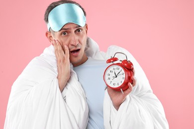 Photo of Overslept man with sleep mask and alarm clock wrapped in blanket on pink background