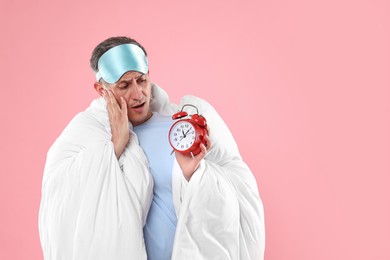 Overslept man with sleep mask and alarm clock wrapped in blanket on pink background, space for text