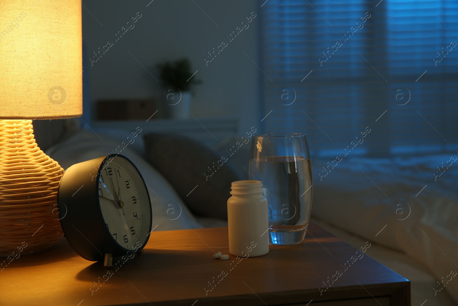 Photo of Insomnia treatment. Glass of water, pills and alarm clock on bedside table in bedroom at night