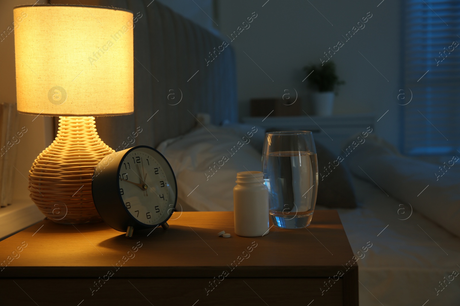 Photo of Insomnia treatment. Glass of water, pills and alarm clock on bedside table in bedroom at night