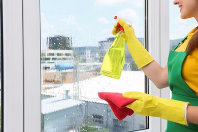 Photo of Housewife with spray bottle of detergent and napkin cleaning window indoors, closeup