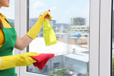 Housewife with spray bottle of detergent and napkin cleaning window indoors, closeup