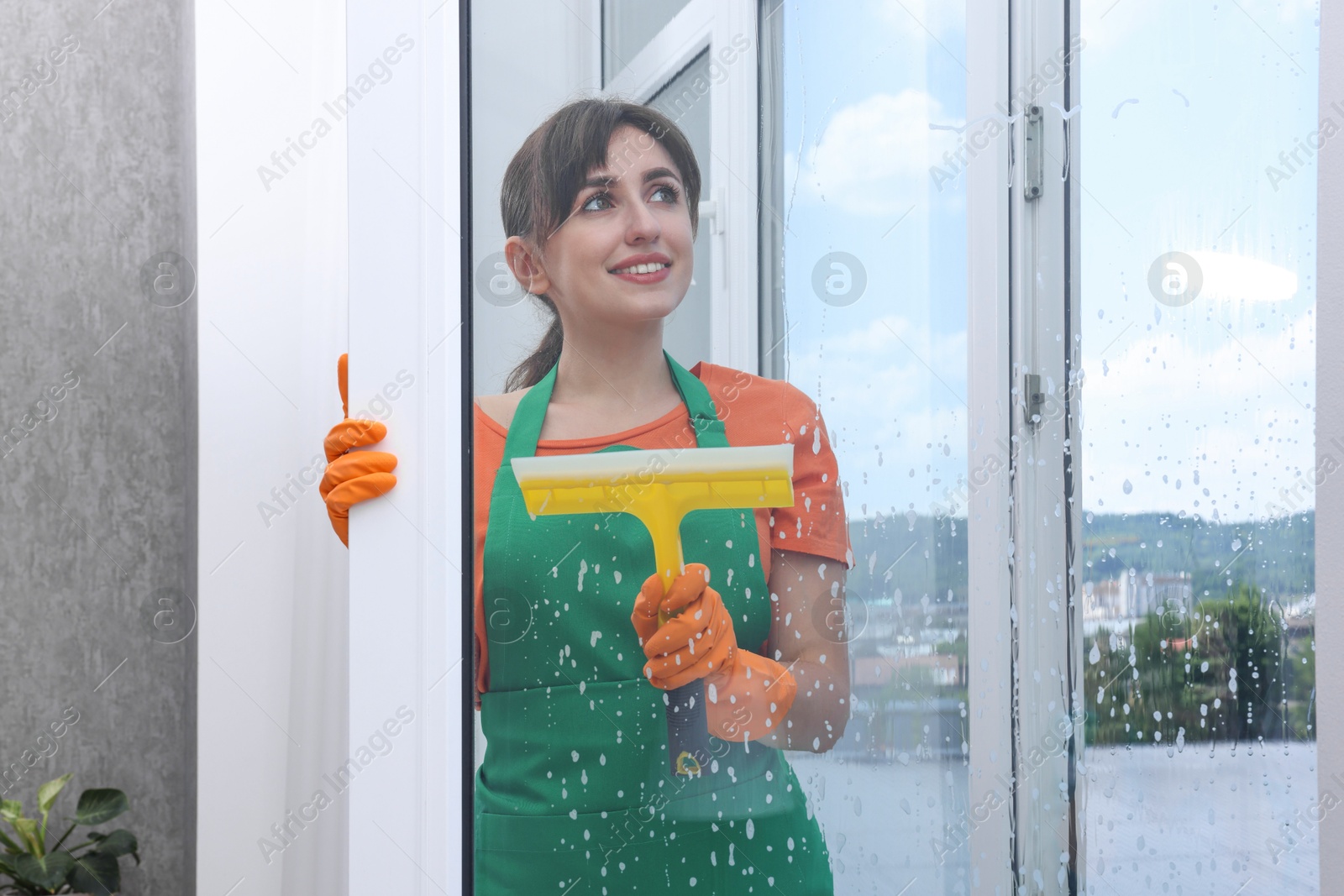 Photo of Young housewife with squeegee cleaning window indoors