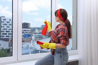 Woman with spray bottle of detergent and napkin cleaning window indoors