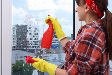 Photo of Woman with spray bottle of detergent and napkin cleaning window indoors, closeup