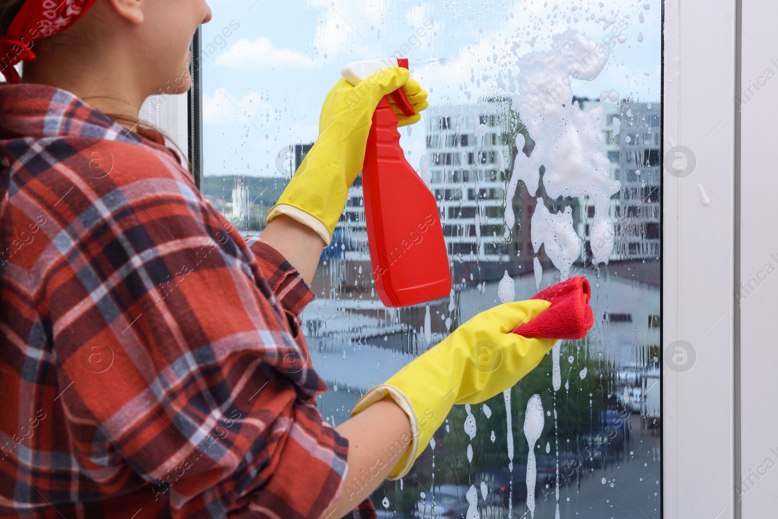 Photo of Woman with spray bottle of detergent and napkin cleaning window indoors, closeup