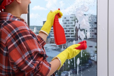 Photo of Woman with spray bottle of detergent and napkin cleaning window indoors, closeup