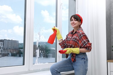 Beautiful young woman with spray bottle of detergent and napkin cleaning window indoors