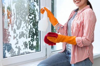 Woman with spray bottle of detergent and napkin cleaning window indoors, closeup