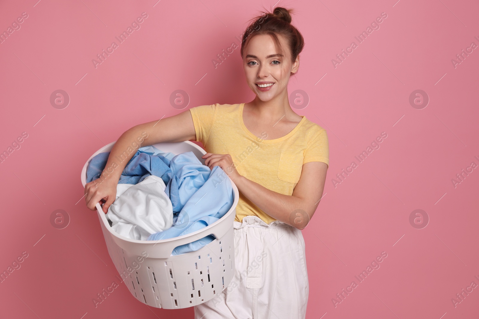 Photo of Happy young housewife with basket full of laundry on pale pink background