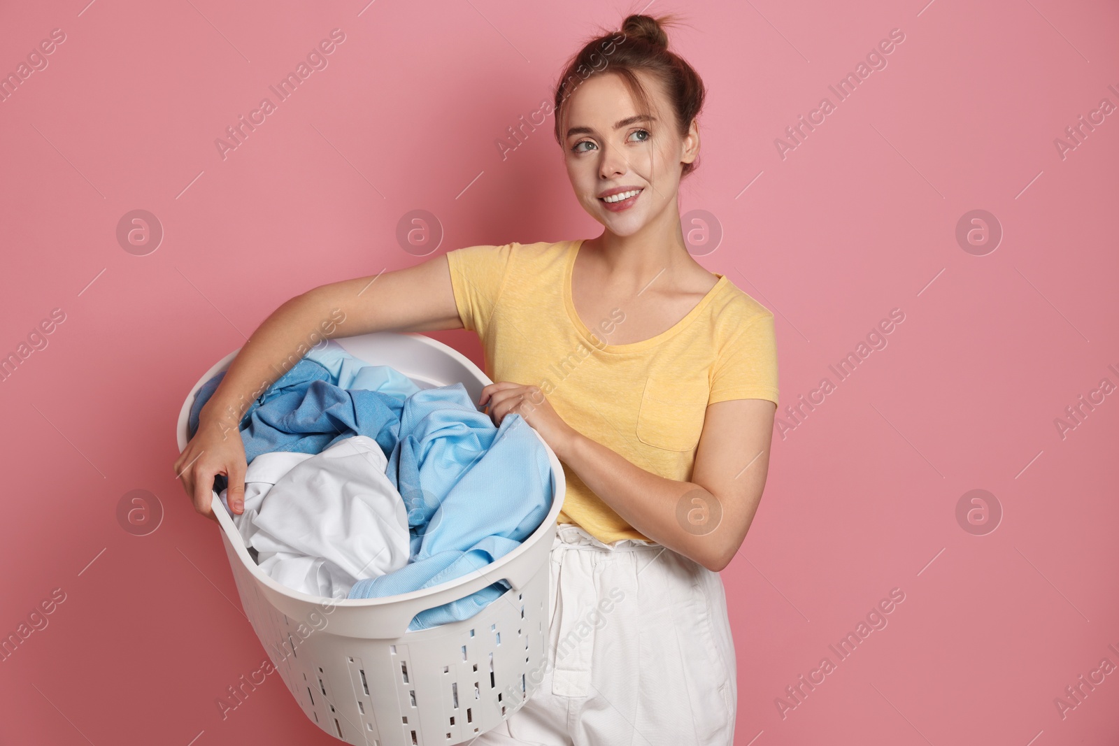 Photo of Happy young housewife with basket full of laundry on pale pink background