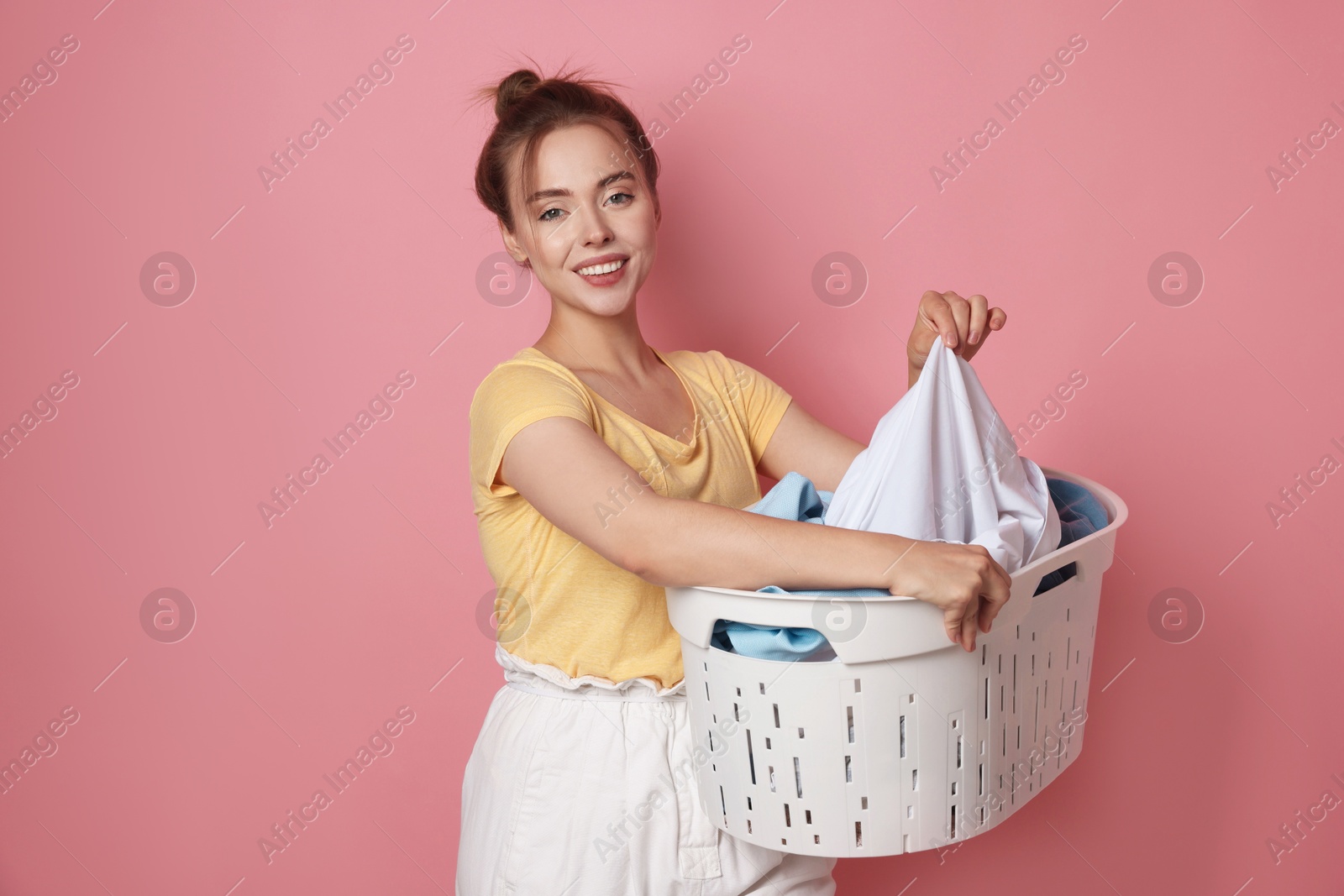 Photo of Happy young housewife with basket full of laundry on pale pink background