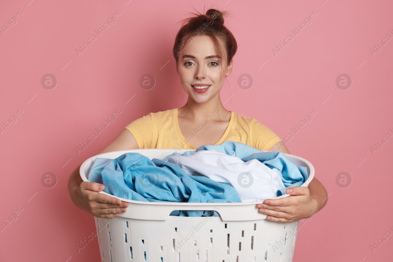 Photo of Happy young housewife with basket full of laundry on pale pink background