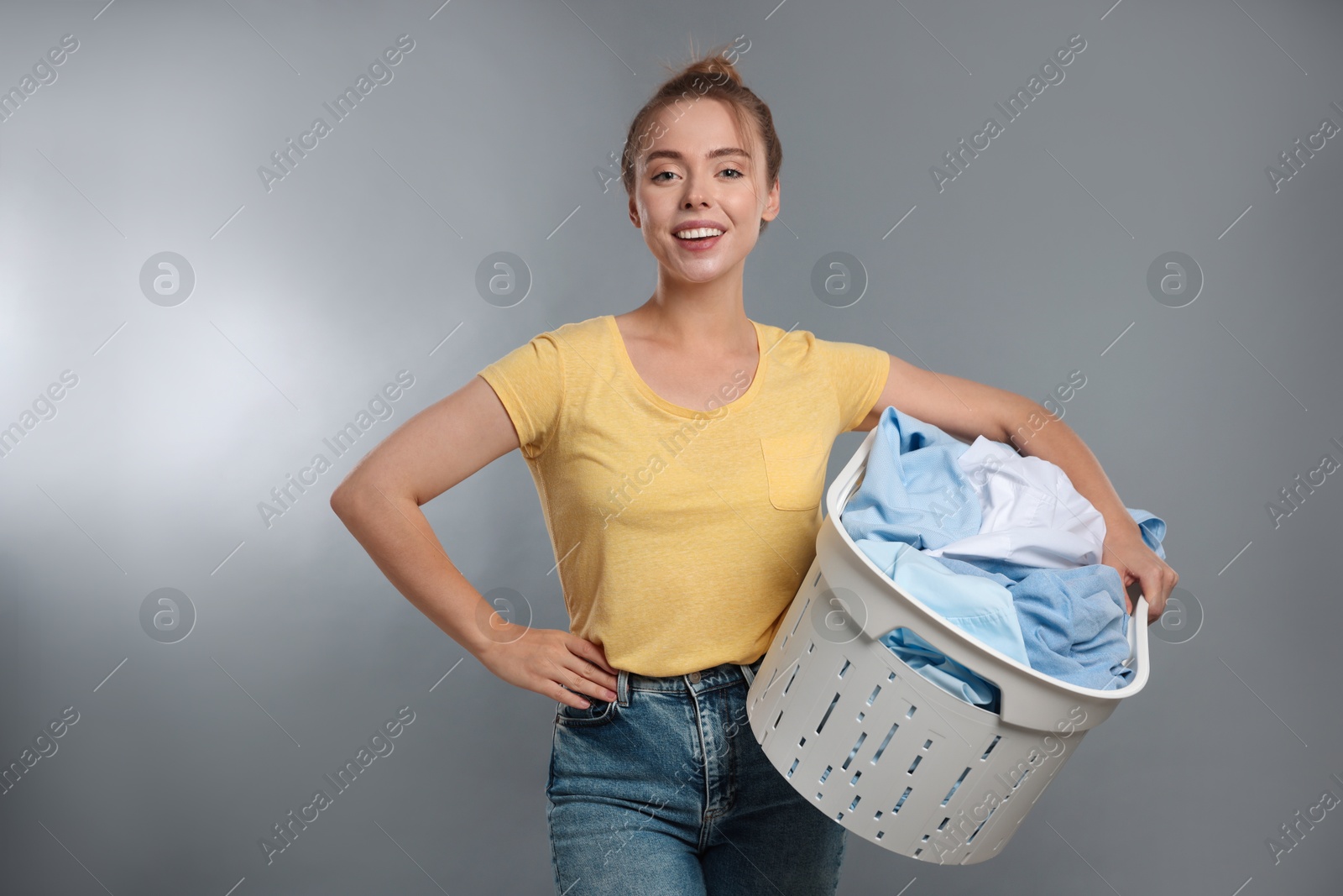 Photo of Happy young housewife with basket full of laundry on grey background