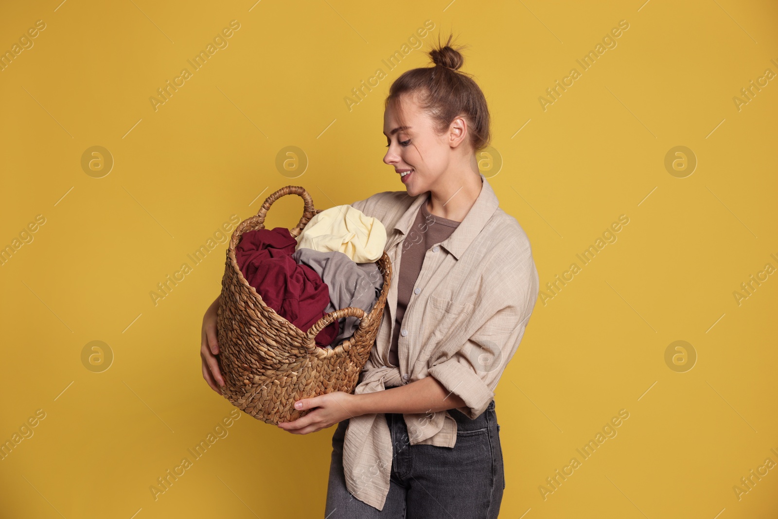 Photo of Happy young housewife with basket full of laundry on yellow background