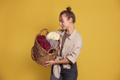 Happy young housewife with basket full of laundry on yellow background