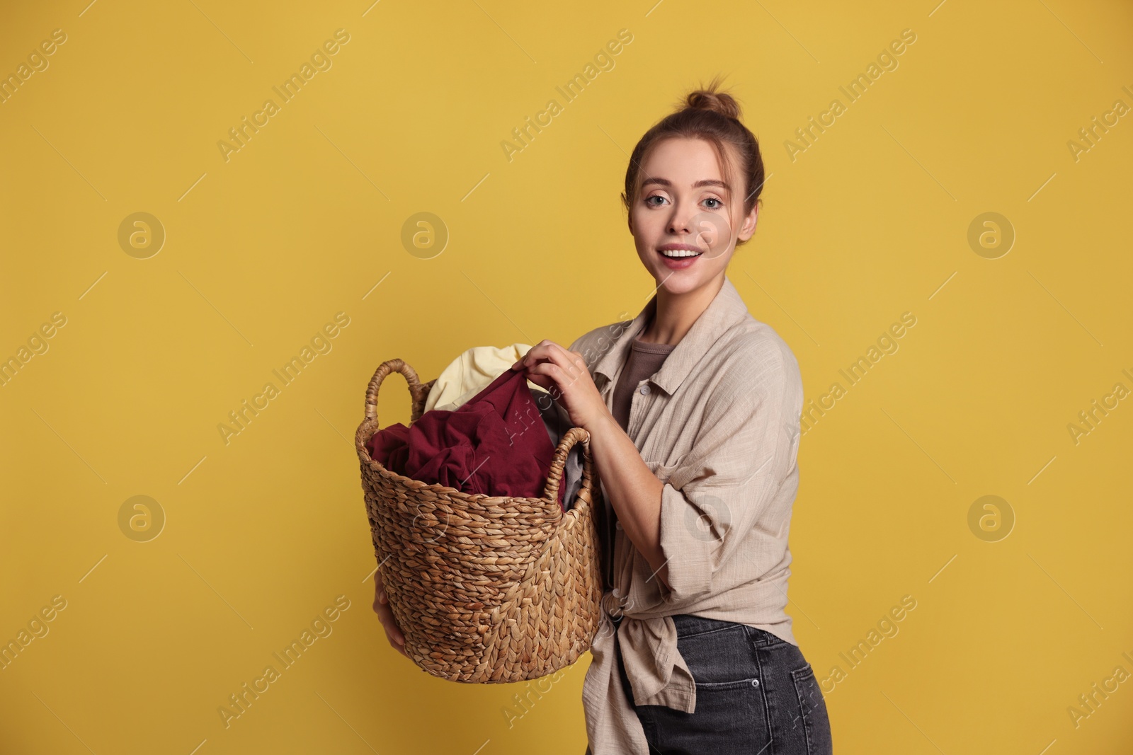 Photo of Happy young housewife with basket full of laundry on yellow background