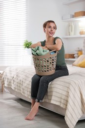 Happy young housewife with basket full of laundry on bed at home