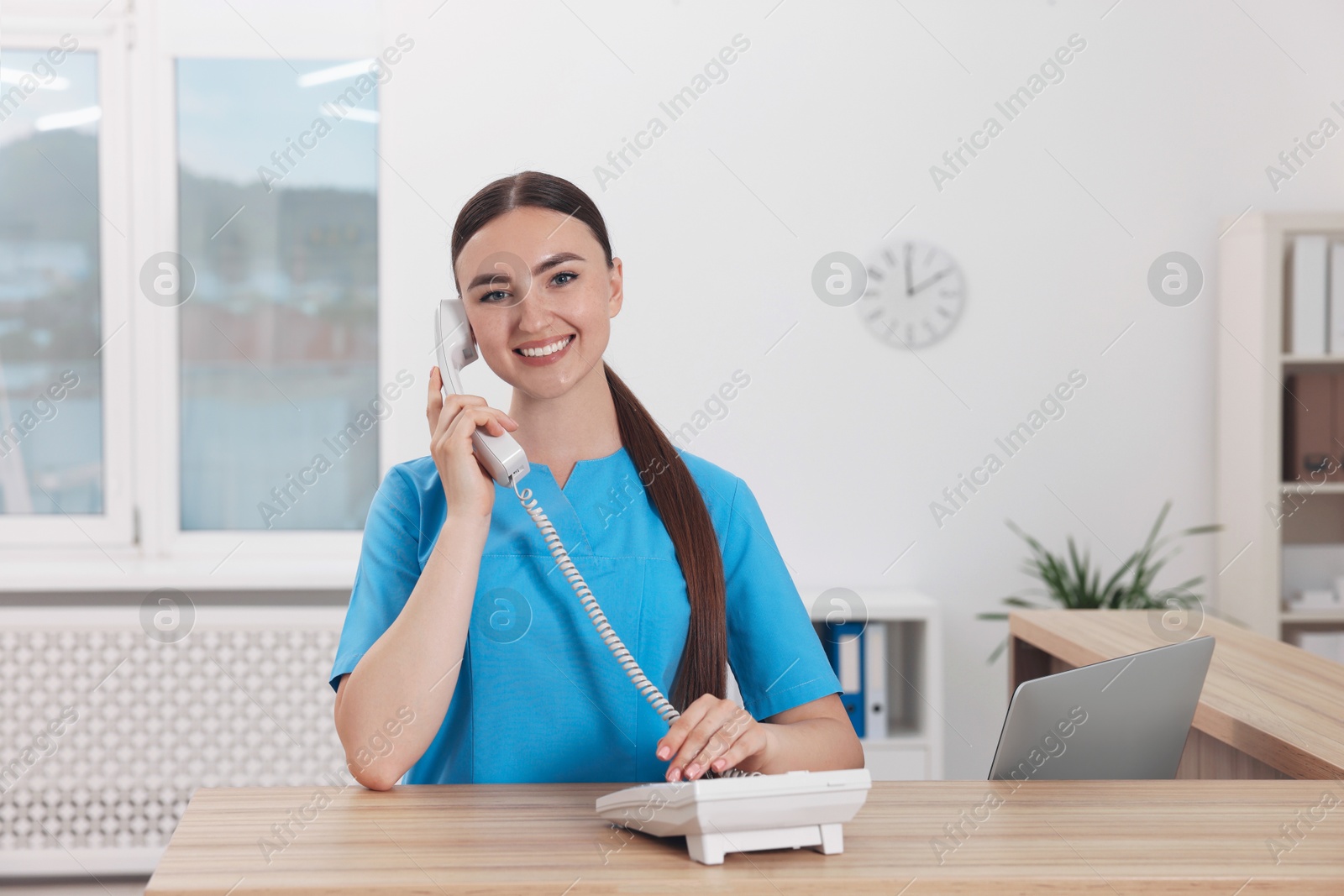 Photo of Professional receptionist talking on phone at wooden desk in hospital