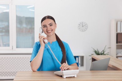 Professional receptionist talking on phone at wooden desk in hospital