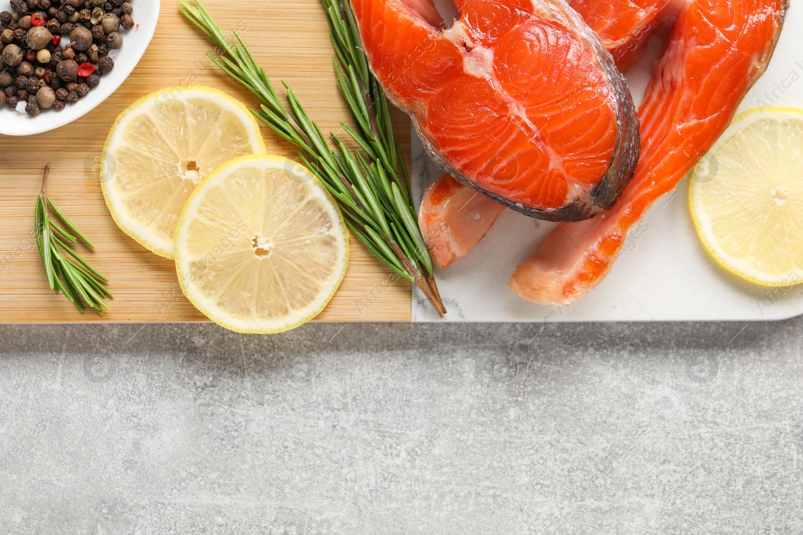 Photo of Board with fresh salmon steaks, peppercorns, rosemary and lemon on grey textured table, top view. Space for text