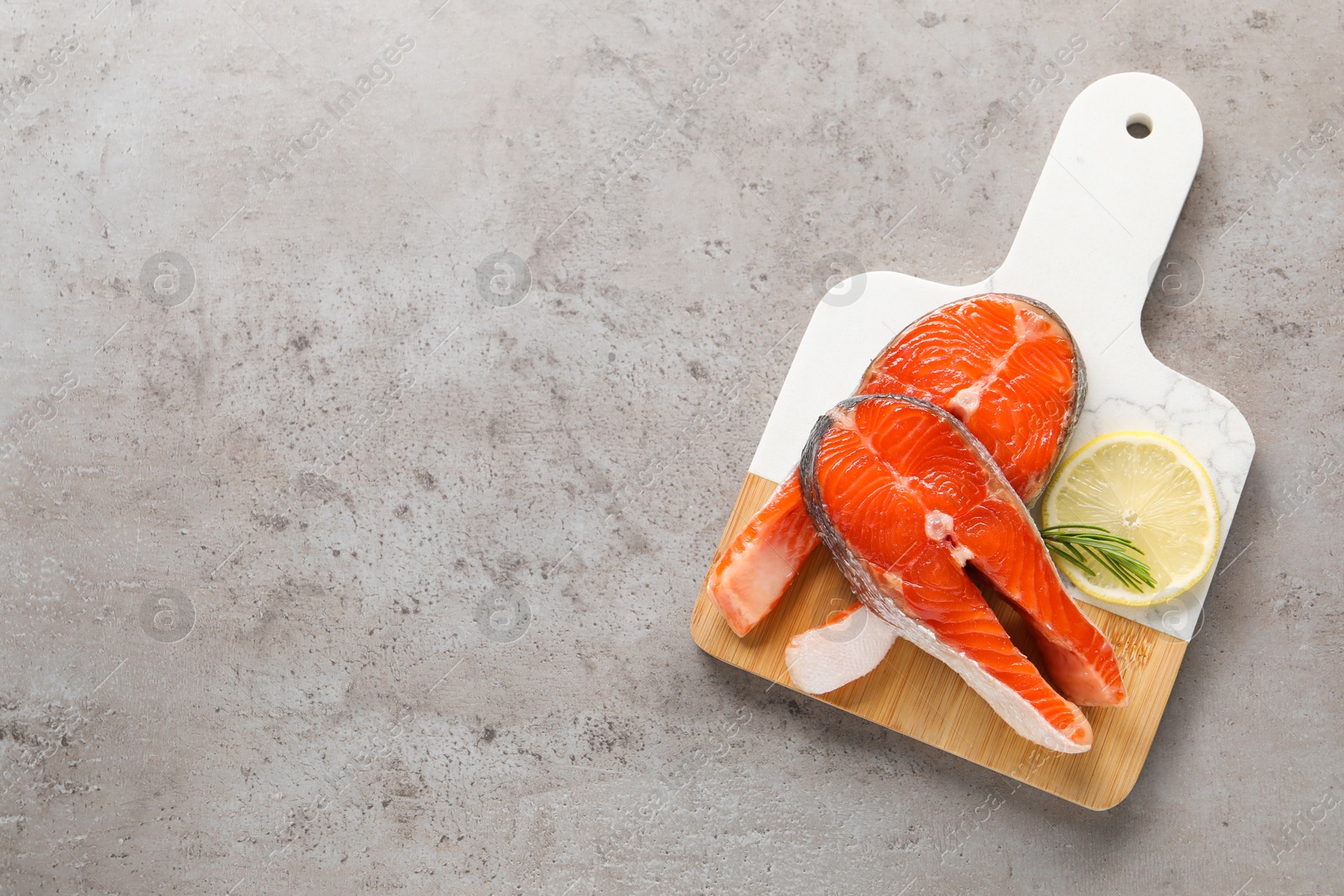 Photo of Board with fresh salmon steaks, rosemary and lemon on grey textured table, top view. Space for text