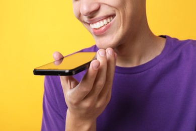 Young man recording voice message via smartphone on yellow background, closeup