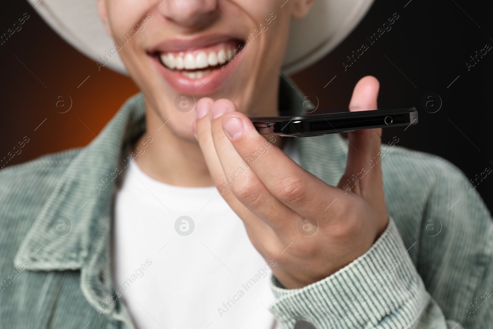 Photo of Young man recording voice message via smartphone on dark background, closeup