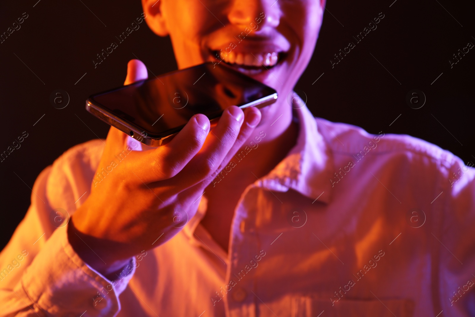 Photo of Young man recording voice message via smartphone on dark background with neon lights, closeup