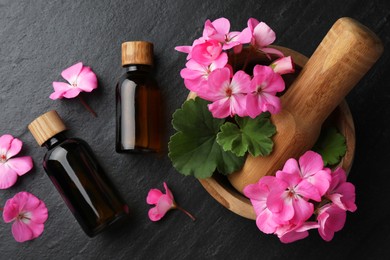 Photo of Bottles of geranium essential oil, beautiful flowers and mortar with pestle on black table, flat lay