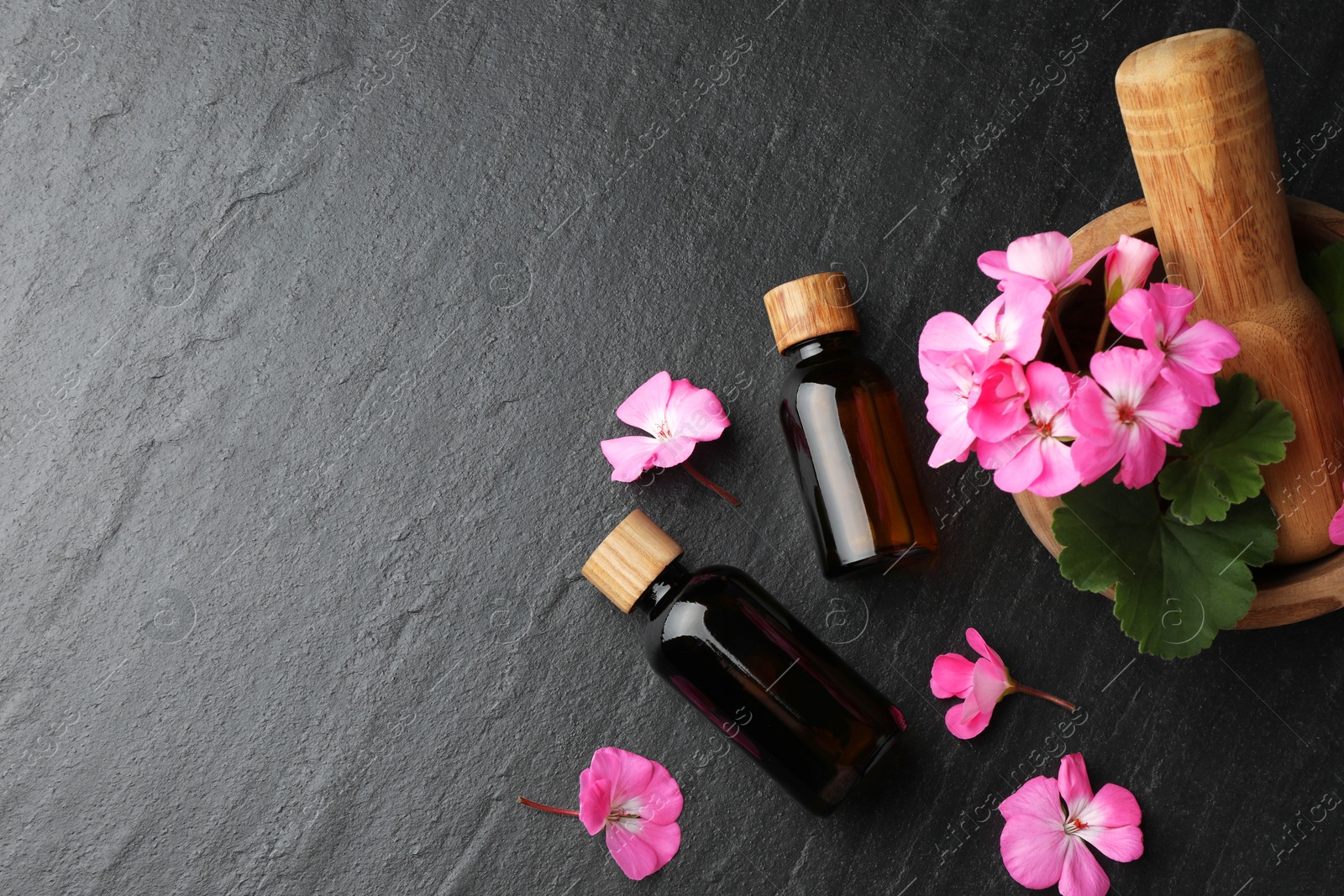 Photo of Bottles of geranium essential oil, beautiful flowers and mortar with pestle on black table, flat lay. Space for text