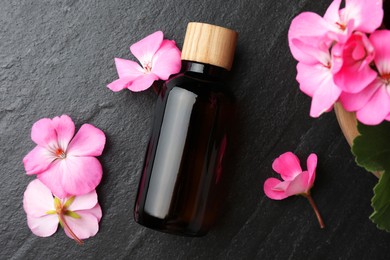 Photo of Bottle of geranium essential oil and beautiful flowers on black table, flat lay