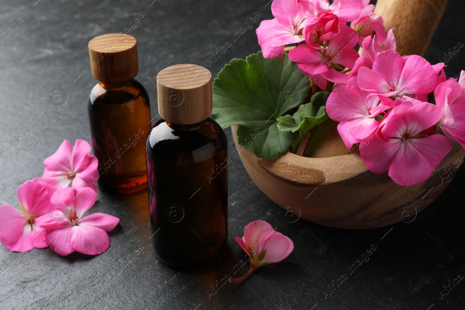 Photo of Bottles of geranium essential oil, beautiful flowers and mortar with pestle on black table, closeup