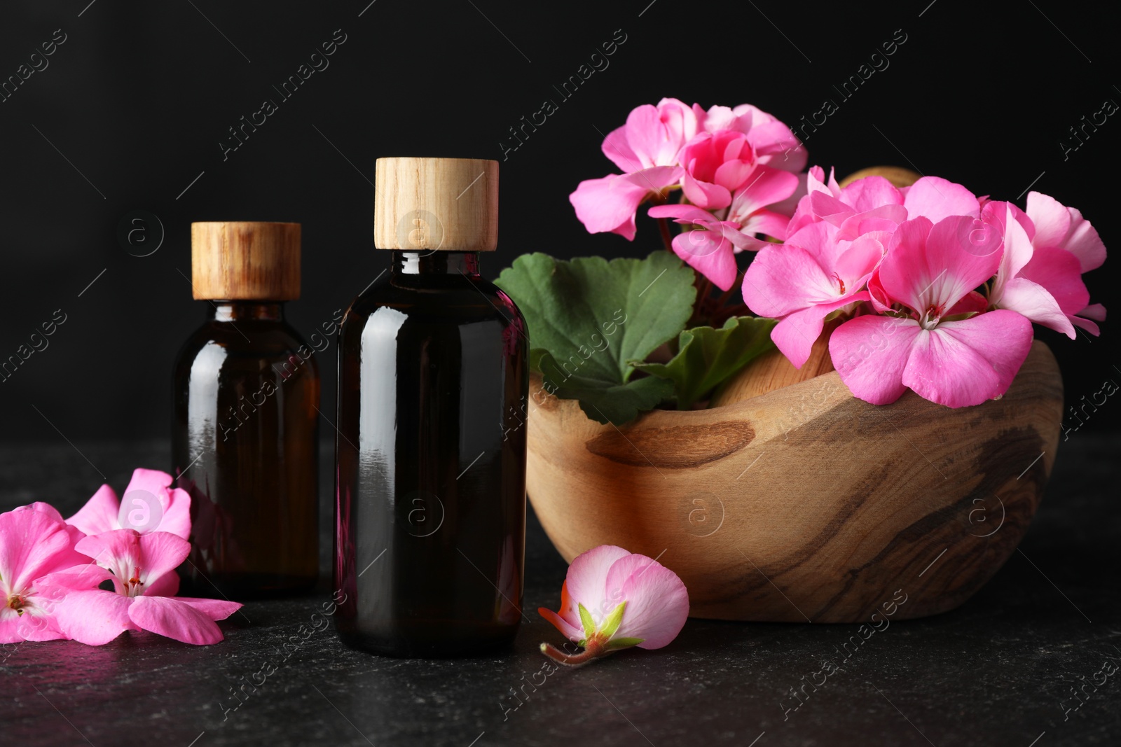 Photo of Bottles of geranium essential oil and beautiful flowers on black table, closeup