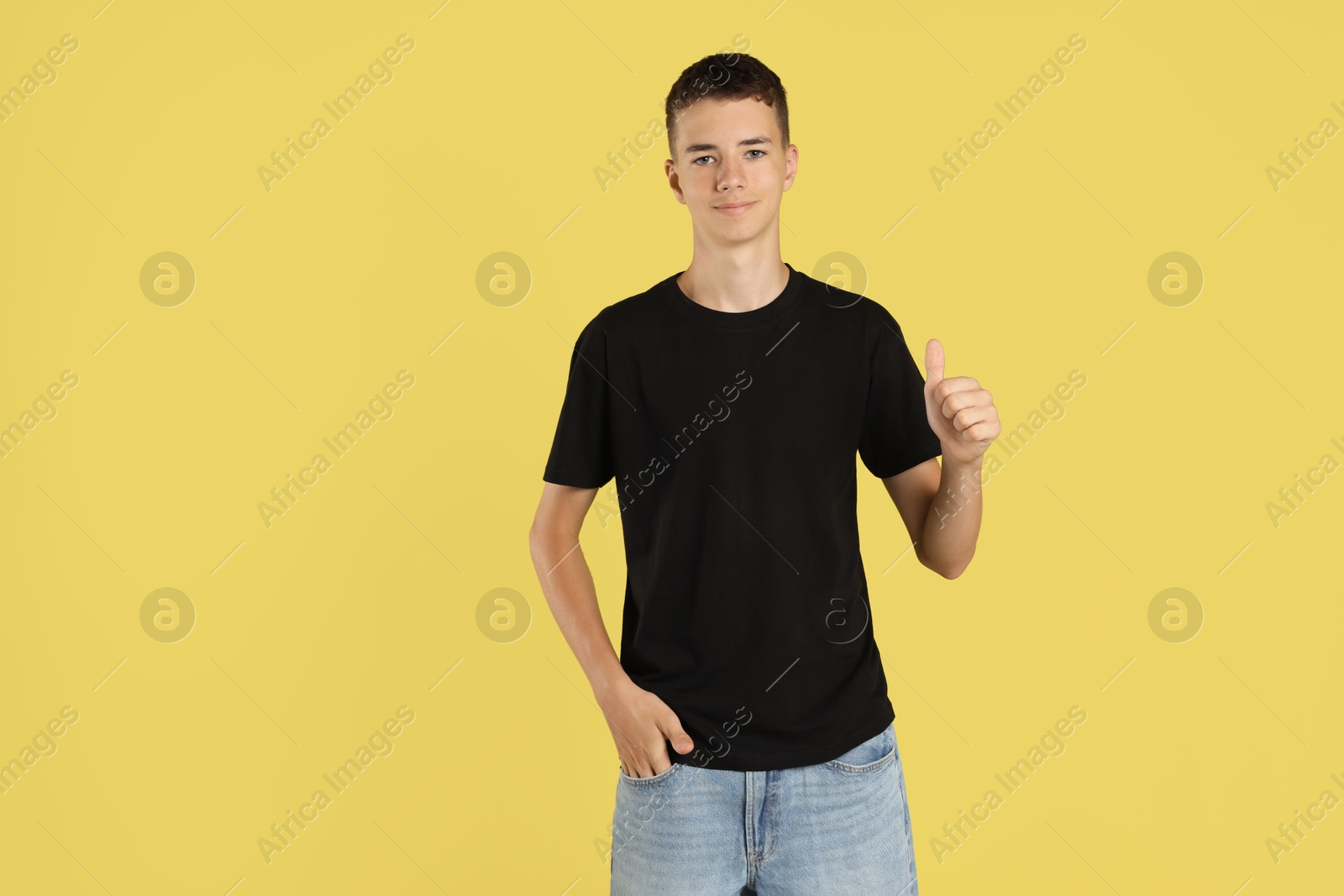 Photo of Teenage boy wearing black t-shirt and showing thumbs up on yellow background, space for text