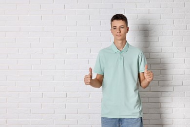 Teenage boy wearing light green t-shirt and showing thumbs up near white brick wall, space for text