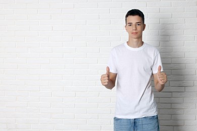 Photo of Teenage boy wearing t-shirt and showing thumbs up near white brick wall, space for text