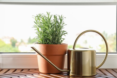 Photo of Aromatic rosemary plant in pot and watering can on wooden table near window indoors